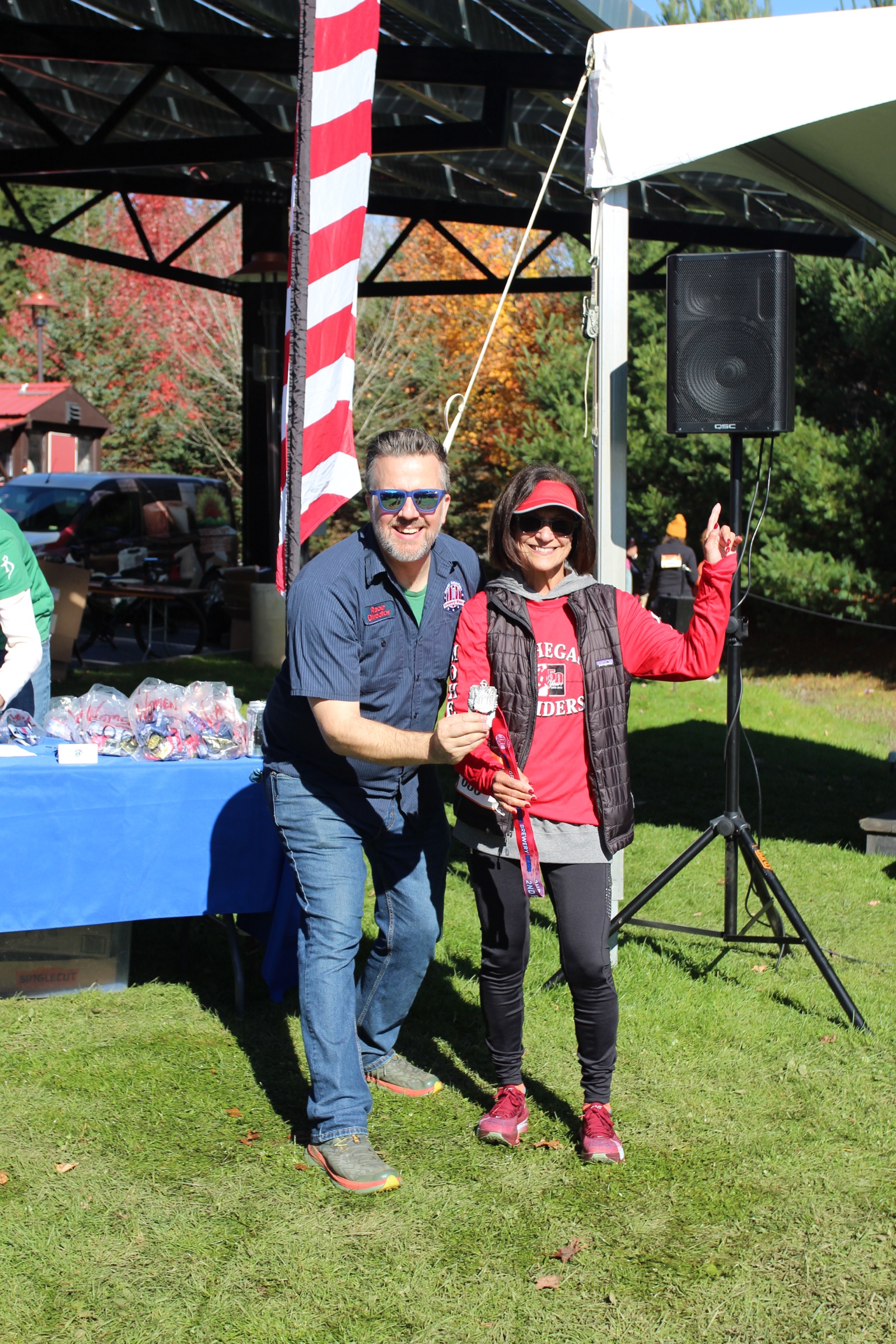 A race director poses with a smiling medal recipient beside an awards table.