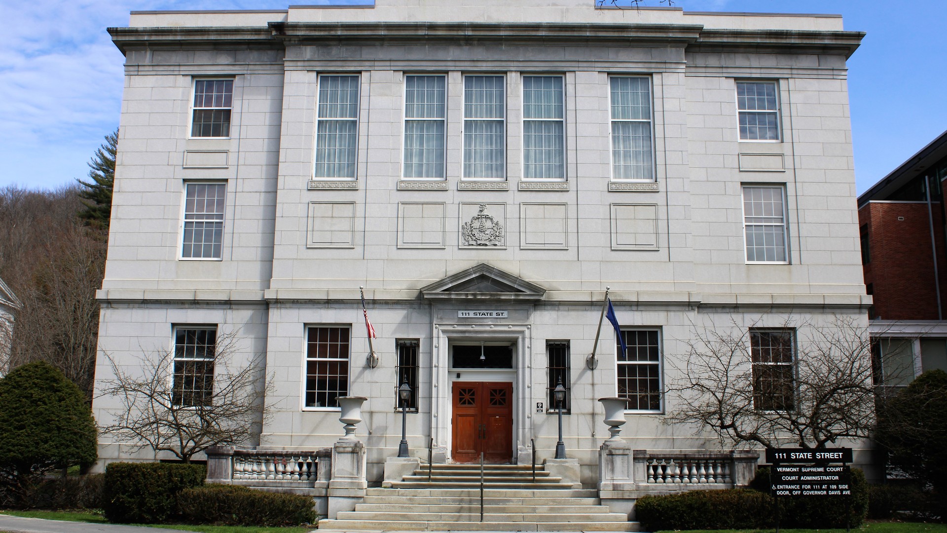 A three-story white stone building with tall windows, a red wooden door, two flags and a sign reading 111 State Street stands behind a lawn and a stone walkway.