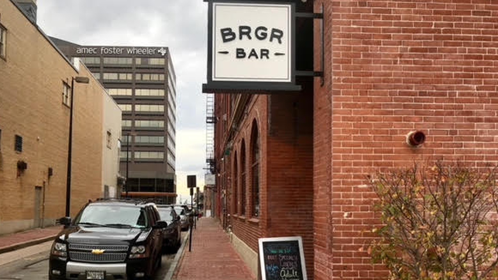 A red brick building corner with a hanging white BRGR BAR sign, an A-frame chalkboard on the sidewalk, and cars parked along Brown Street.