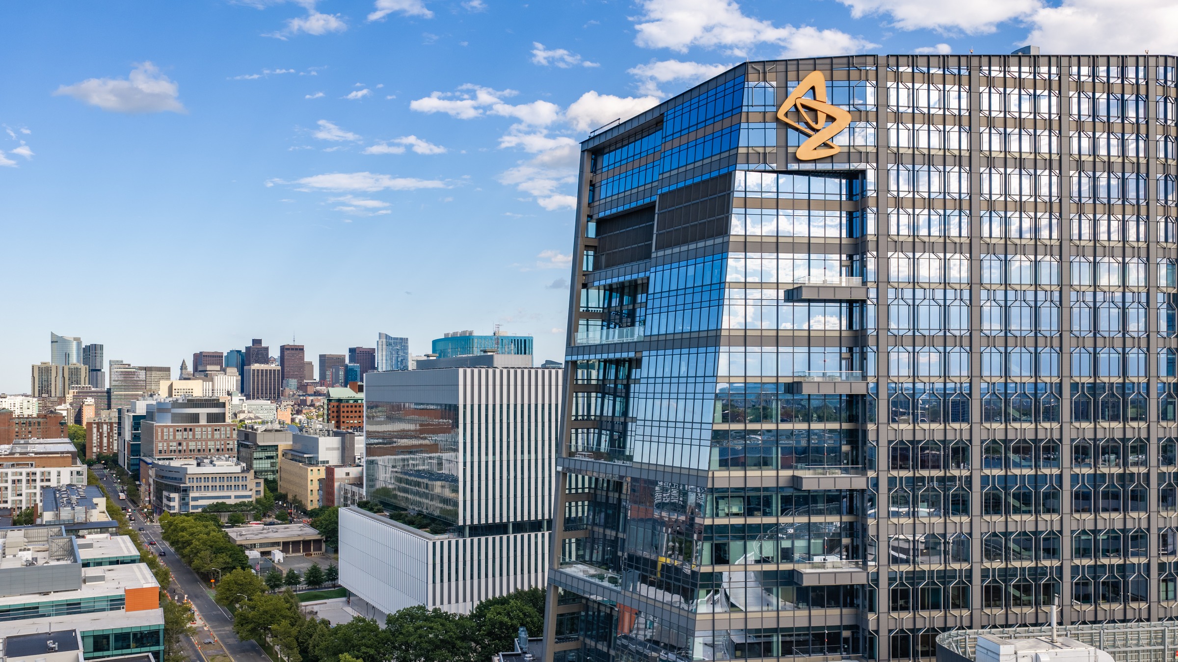 A glass high-rise with a gold AstraZeneca logo stands over Kendall Square streets with the Boston skyline beyond.