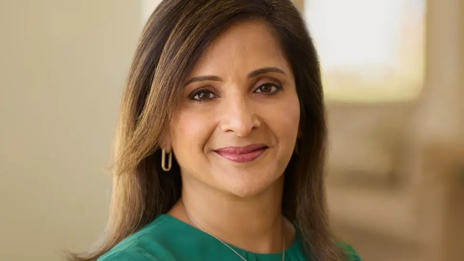 Yamini Rangan smiles at the camera in a green round-neck blouse, framed from the upper chest up against a soft blurred beige background.