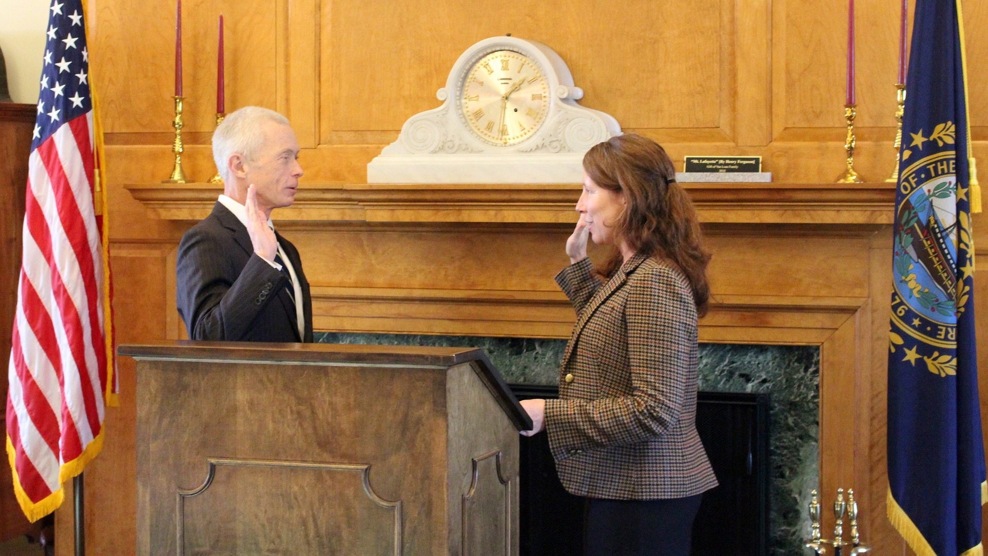 A gray-haired man in a dark suit and a woman in a checked jacket each raise a right hand in a wood-paneled room with a fireplace, a mantel clock and U.S. and New Hampshire flags.