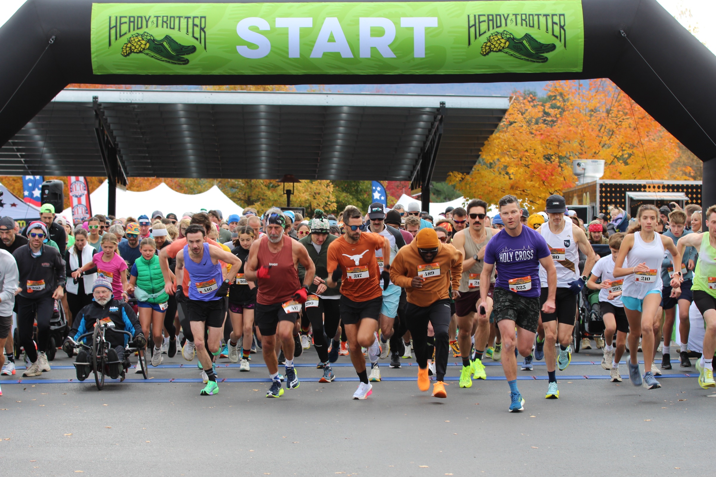 Runners, including a participant on a hand cycle, cross the start line under a green Heady Trotter arch, with fall foliage behind them.