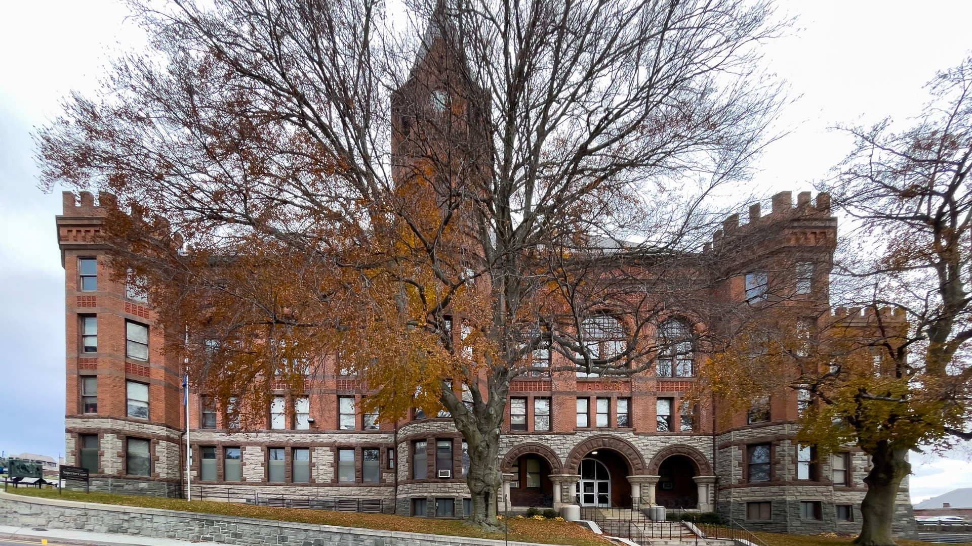 A red brick courthouse with a central tower, crenellated corner towers and arched entrances stands behind a large autumn tree.