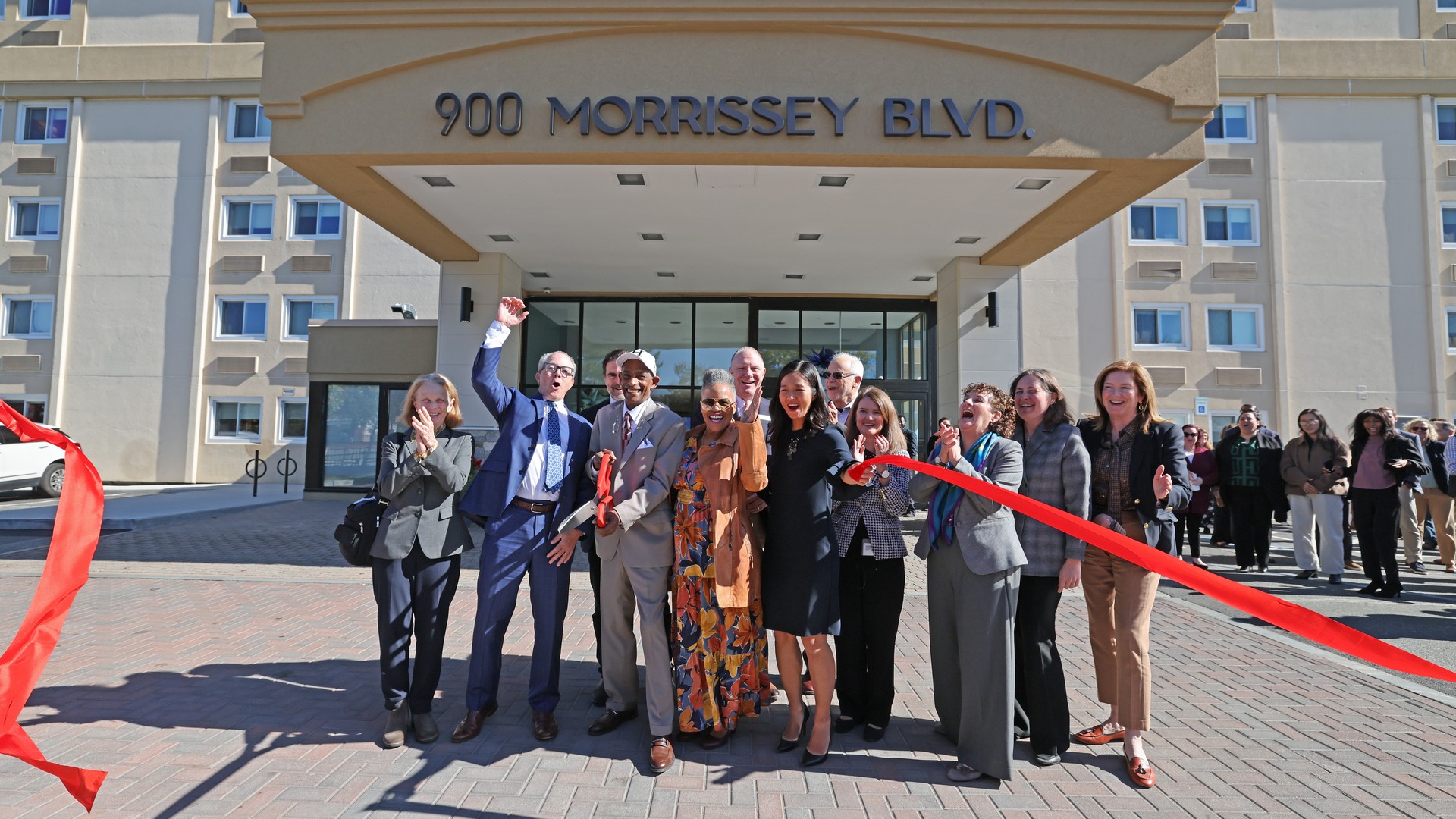 A group of people, one waving and others clapping, stand with streaming red ribbon under an entrance canopy marked 900 Morrissey Blvd. in front of a beige building.