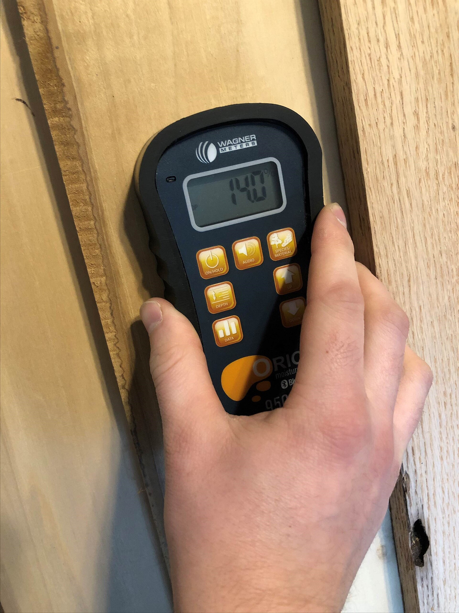 A handheld moisture meter pressed against wood shows a reading of 14 percent.