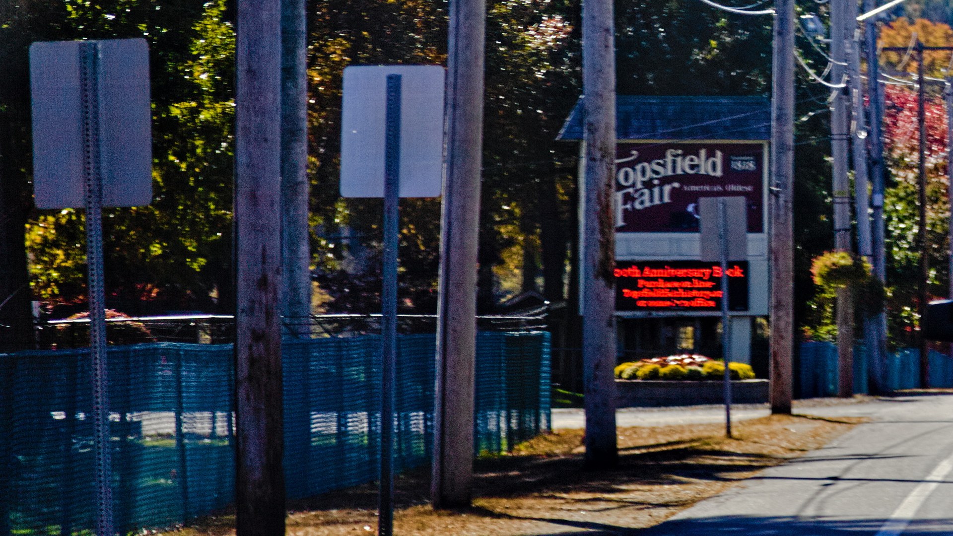 The Topsfield Fair entrance sign beside the road, with a blue construction fence and utility poles in front.