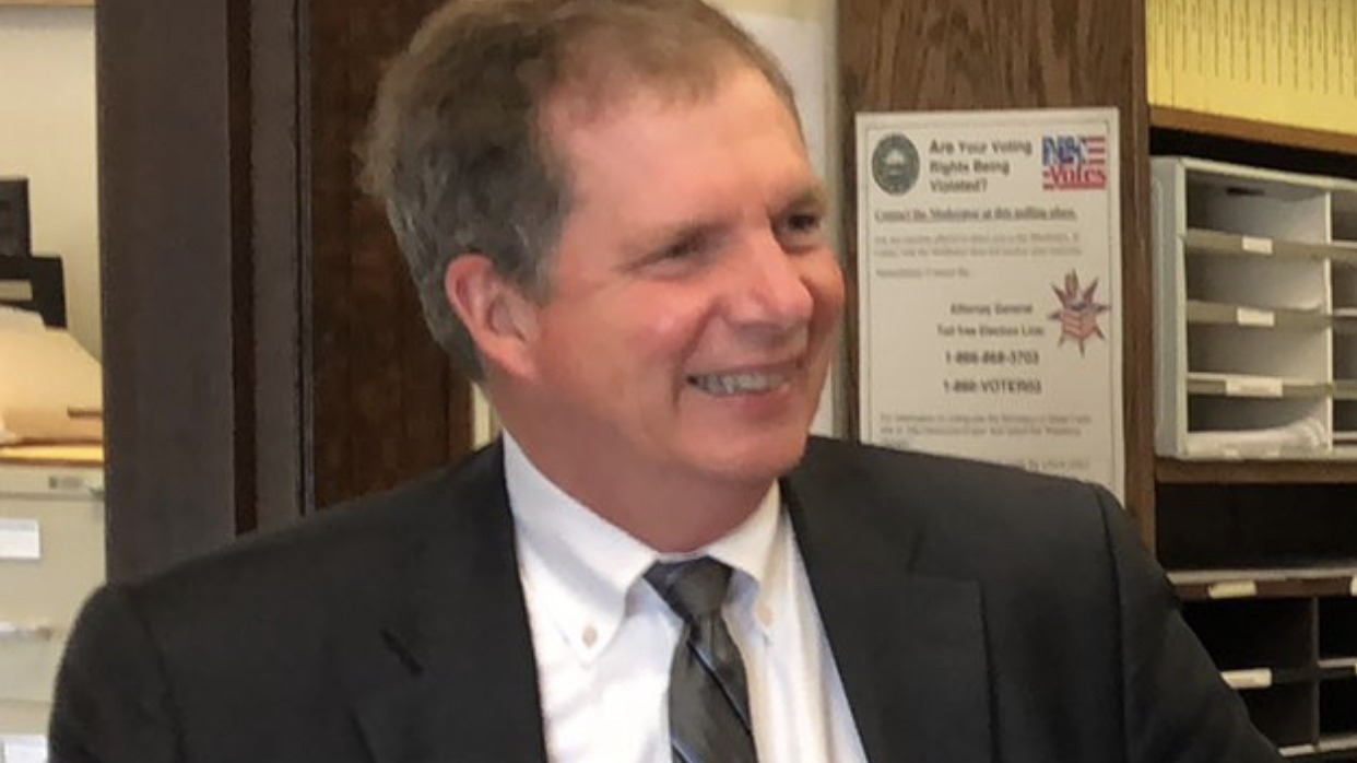 David Scanlan smiles in a dark suit beside a New Hampshire poster that asks "Are Your Voting Rights Being Violated?"