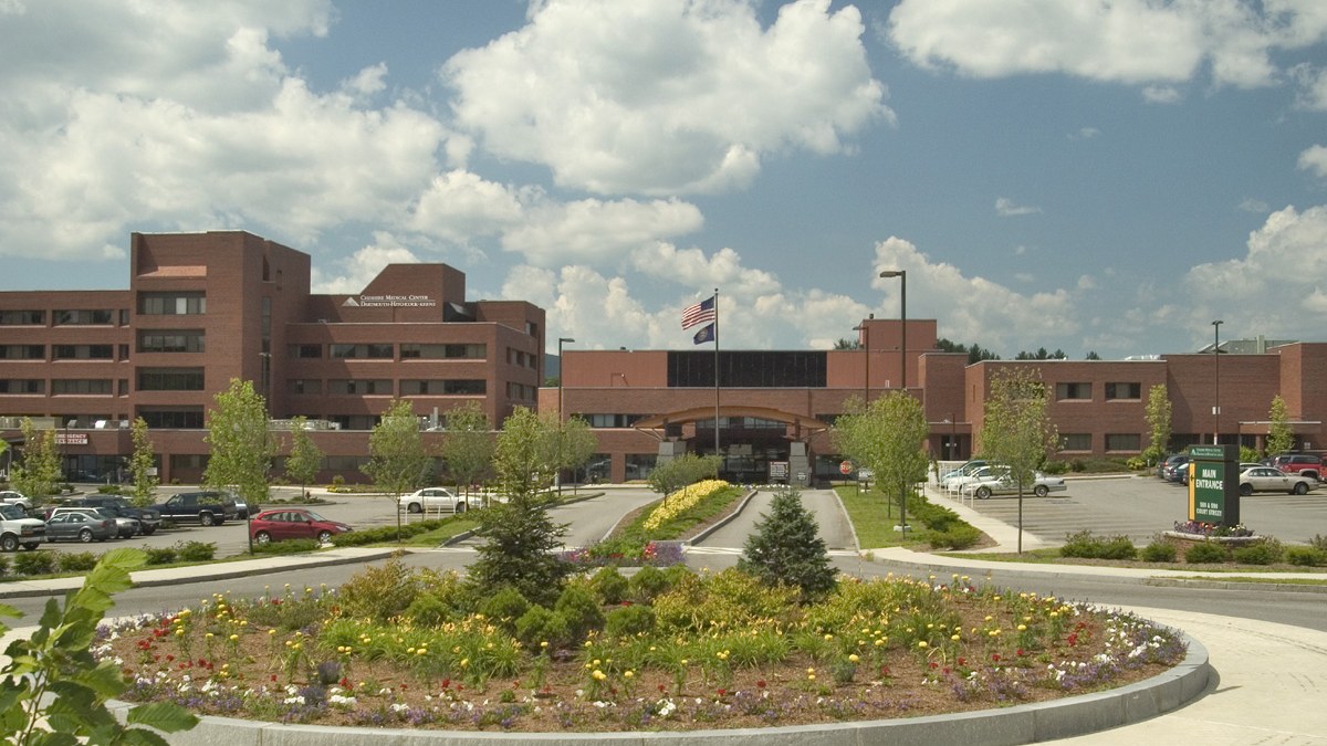 A large brick hospital complex behind a parking lot and a circular flower bed, under a partly cloudy sky.