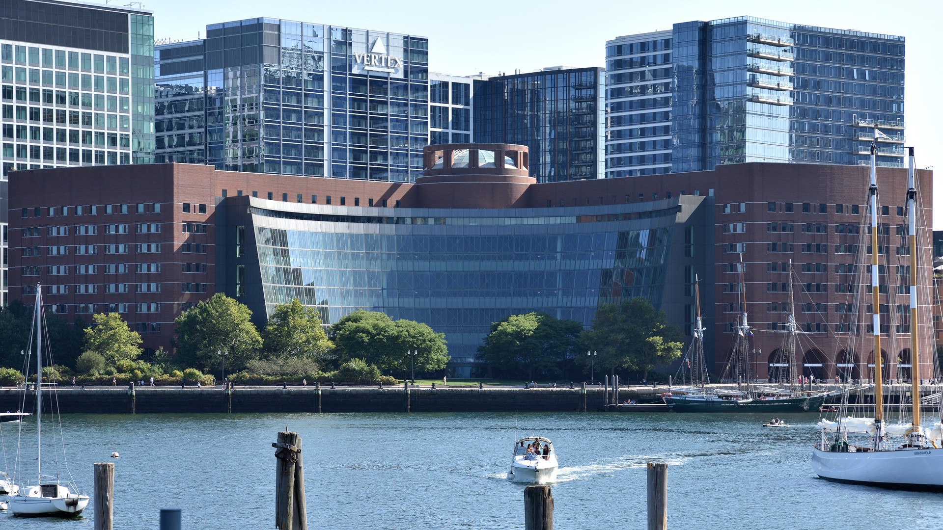 A curved brick and glass building sits on a waterfront in front of glass office towers, with sailboats and a small motorboat on the harbor.