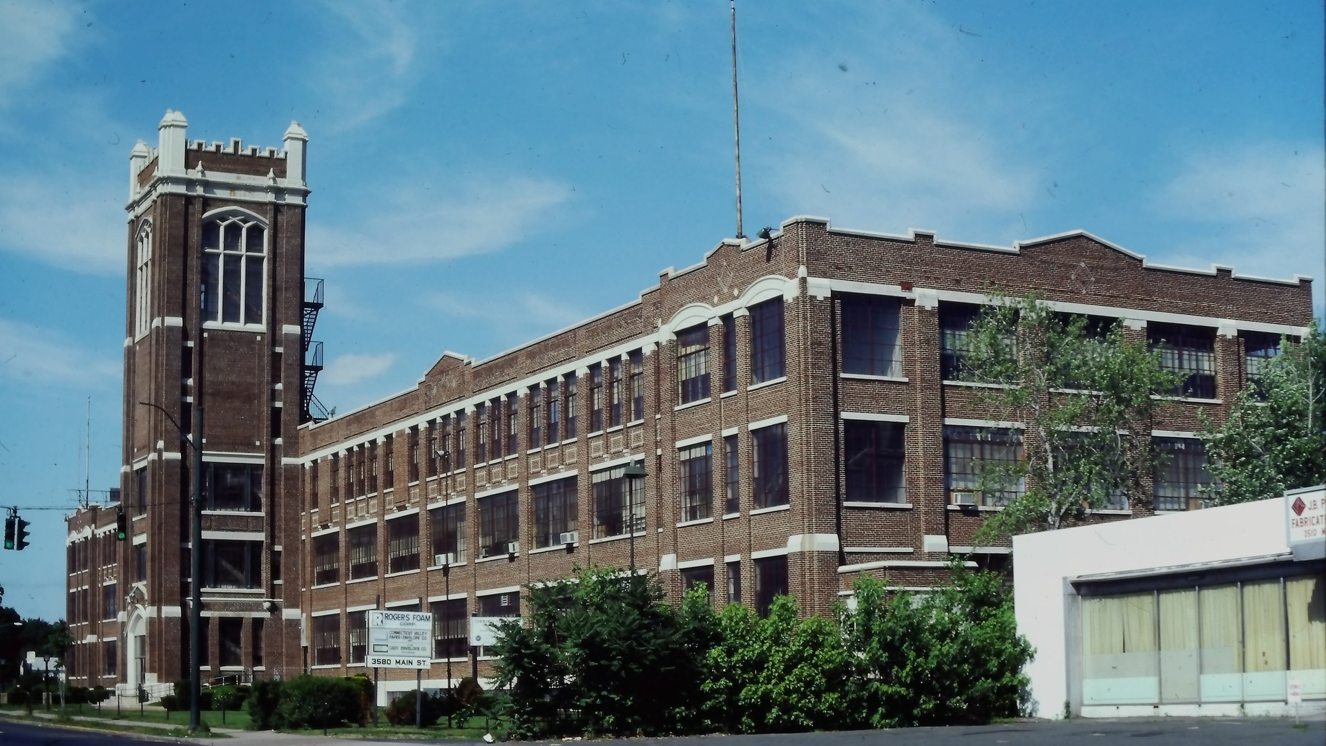 A long brick factory building with a tall square tower at one end, trees along the street, and a low white building in front.