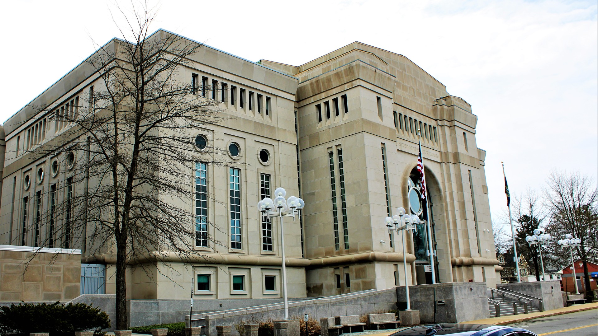 A large tan stone courthouse with tall narrow windows, round windows and an arched entrance stands behind a bare tree and globe lampposts.