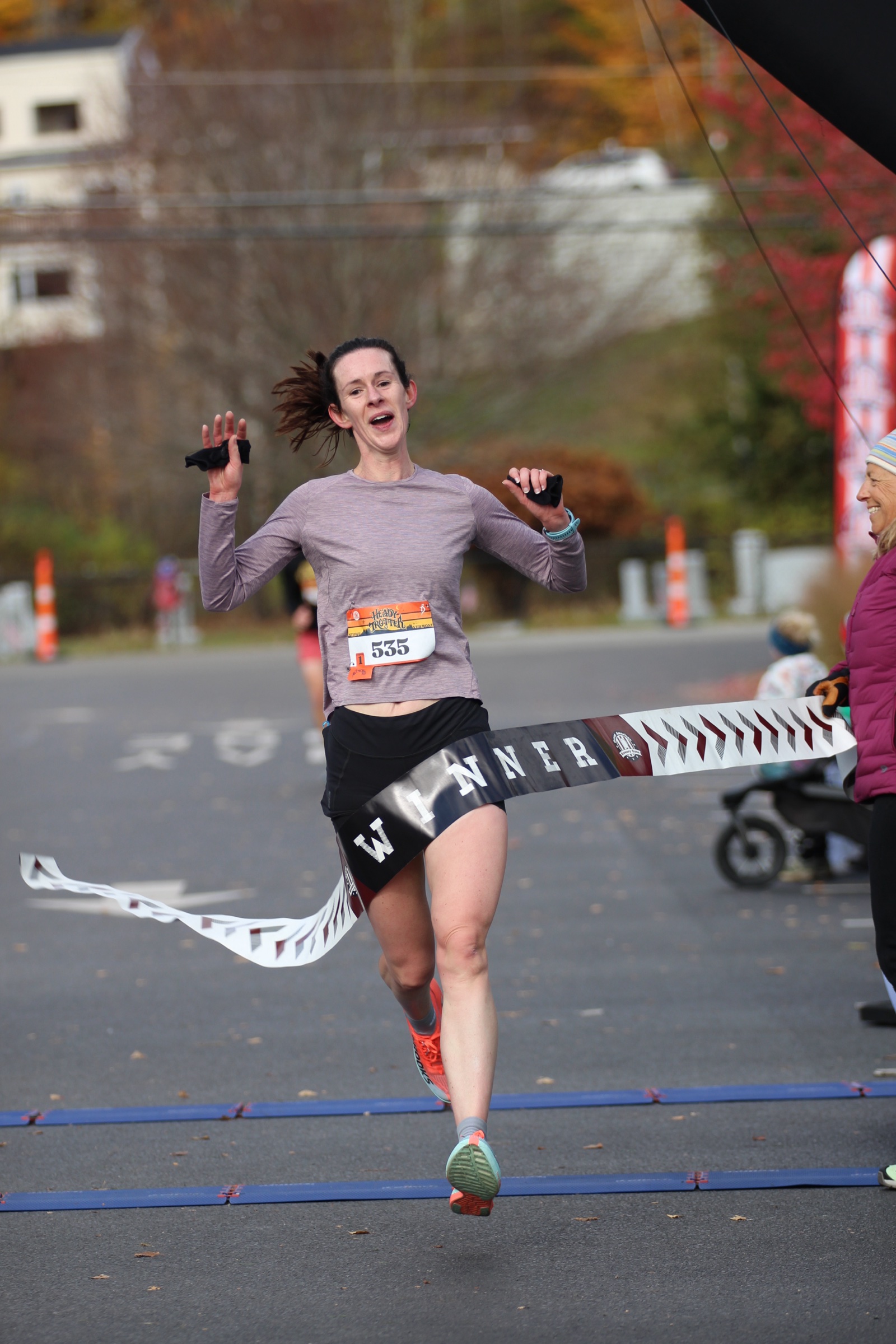 Lauren Hoskin, wearing bib 535 and a lavender long-sleeve shirt, runs through the winner's tape at the Heady Trotter finish.