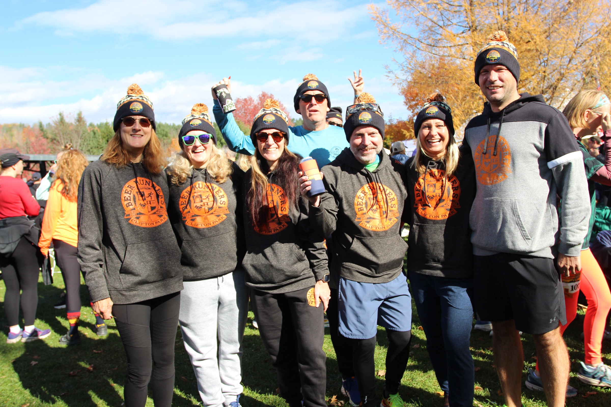 A group of runners in matching gray sweatshirts and knit hats poses on a sunny lawn at the post-race party.